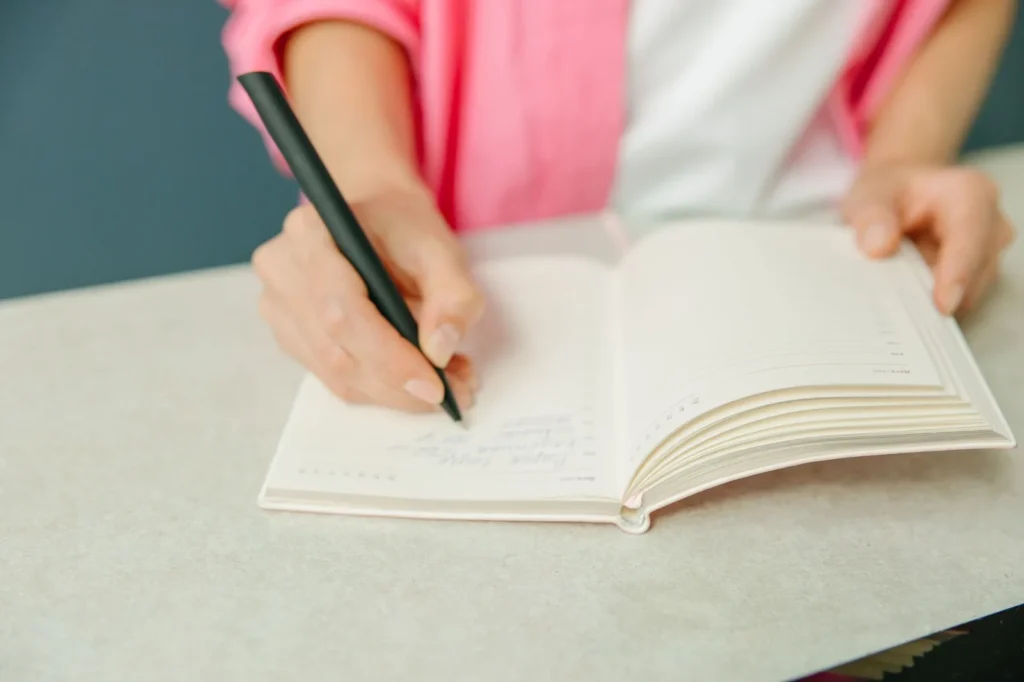 Iemand schrijft met een zwarte pen een planning in een open notitieboek op tafel