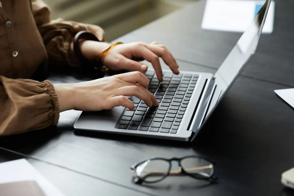 Handen typen op een laptop aan een donkere tafel, met een bril en papieren ernaast