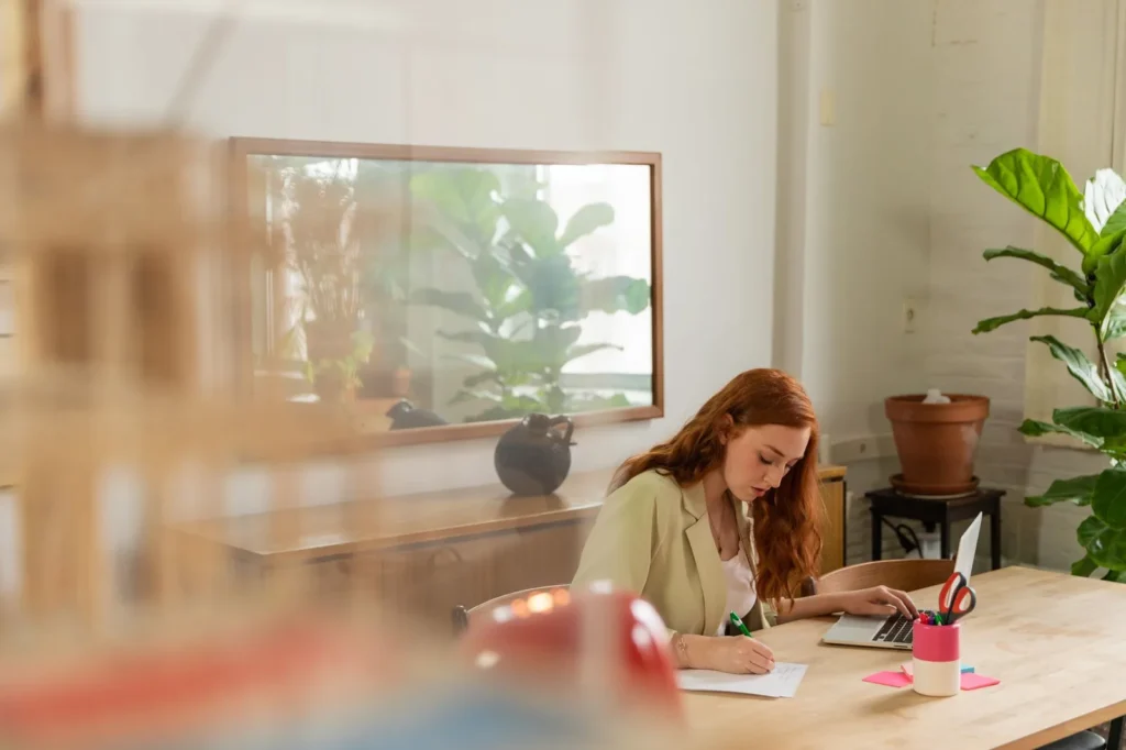Een vrouw schrijft aantekeningen op papier naast haar laptop aan een houten tafel in een licht kantoor met planten