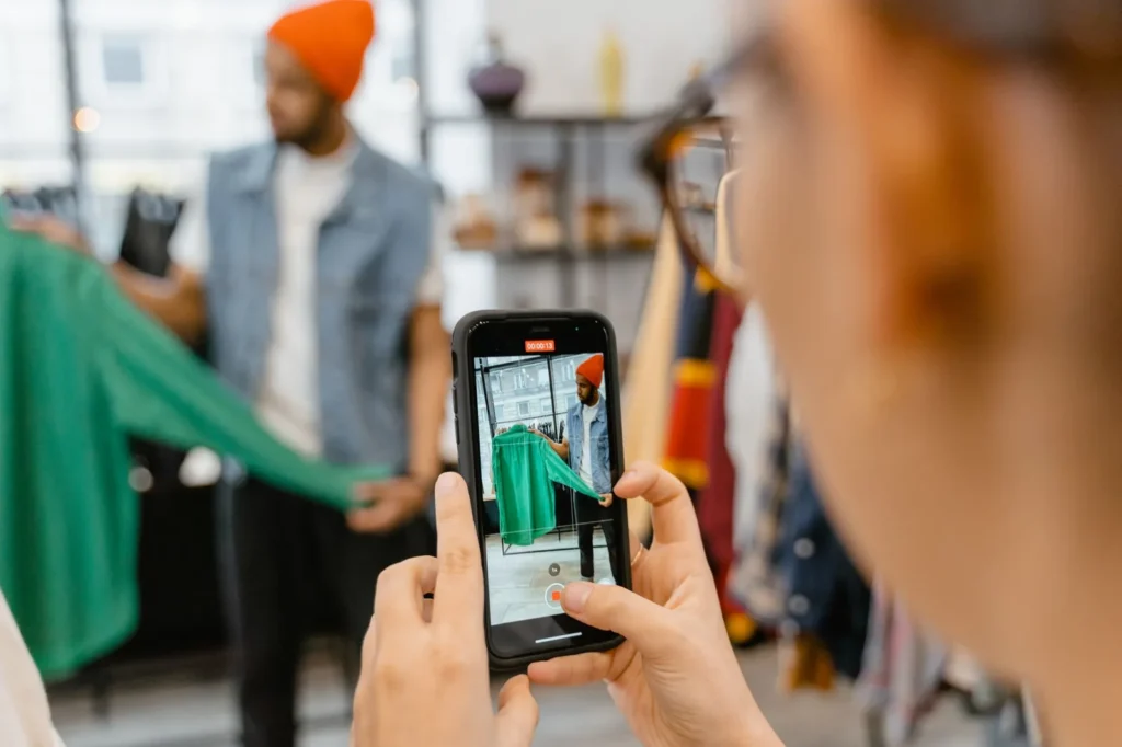 Iemand fotografeert met een smartphone een groen kledingstuk dat een man in een kledingwinkel omhoog houdt