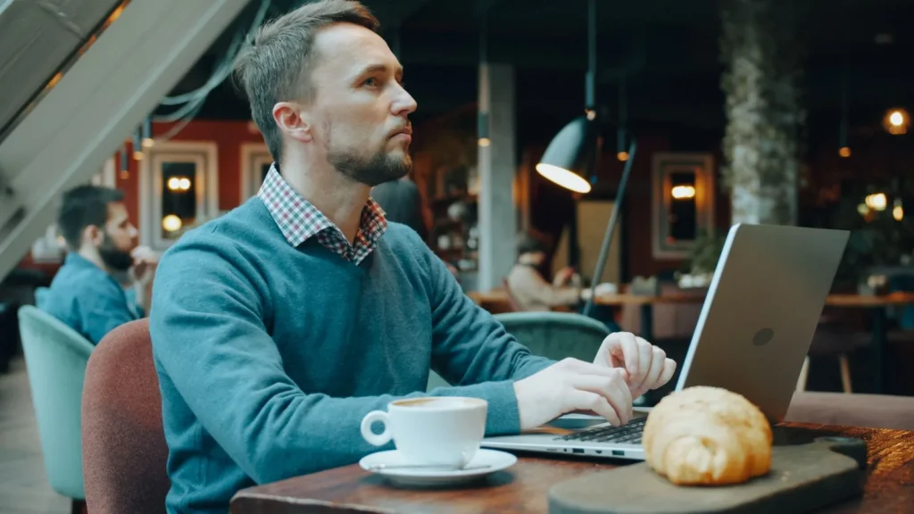Ondernemer werkt aan zijn laptop in een café, met een kop koffie en een croissant op tafel