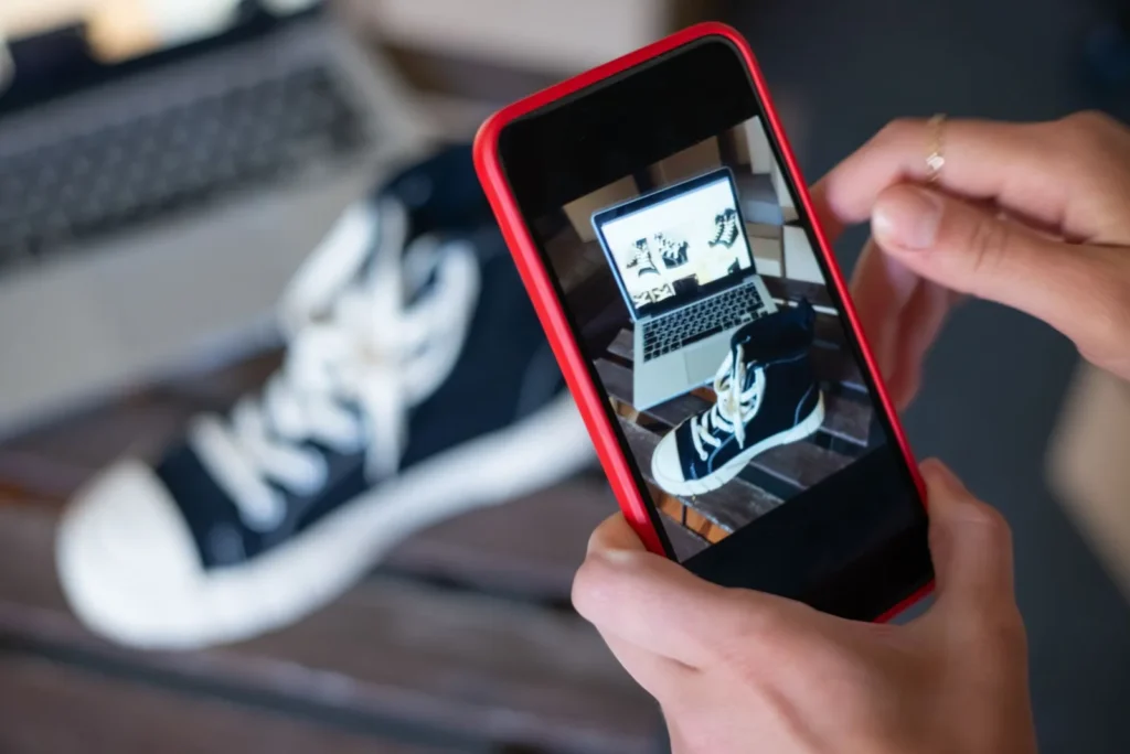 Iemand fotografeert een zwarte sneaker met een smartphone voor een social media post, met een laptop op de achtergrond
