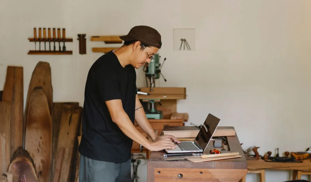Een houtbewerker typt op een laptop aan de werkbank in zijn werkplaats