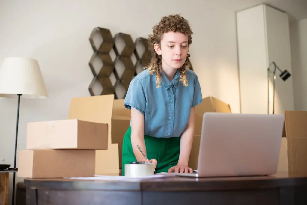 Een jonge ondernemer staat tussen kartonnen verzenddozen aan een bureau en kijkt geconcentreerd naar haar laptop