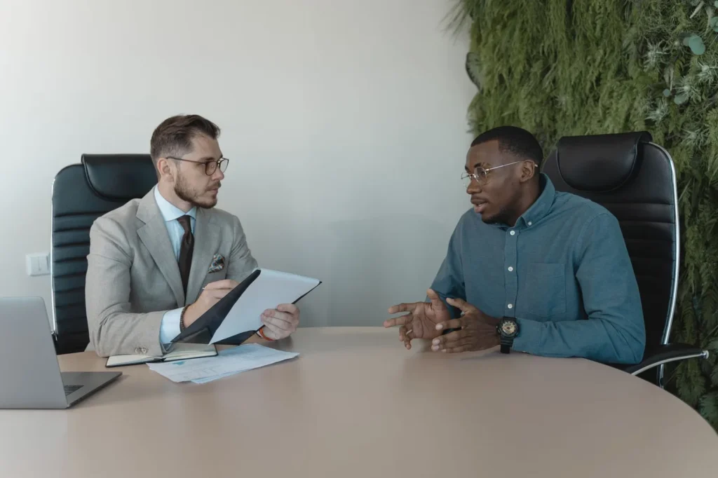 Twee mannen voeren een gesprek aan een kantoortafel, de een maakt aantekeningen op een klembord