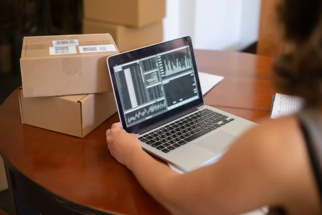 Een ondernemer bekijkt grafieken op een laptop aan een houten tafel met verzenddozen ernaast