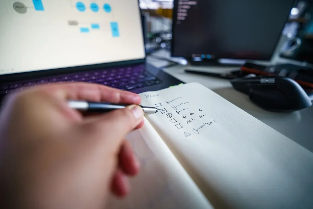 Iemand schrijft een takenlijst met vinkjes in een notitieboek, met op de laptop erachter een schema van gekoppelde stappen