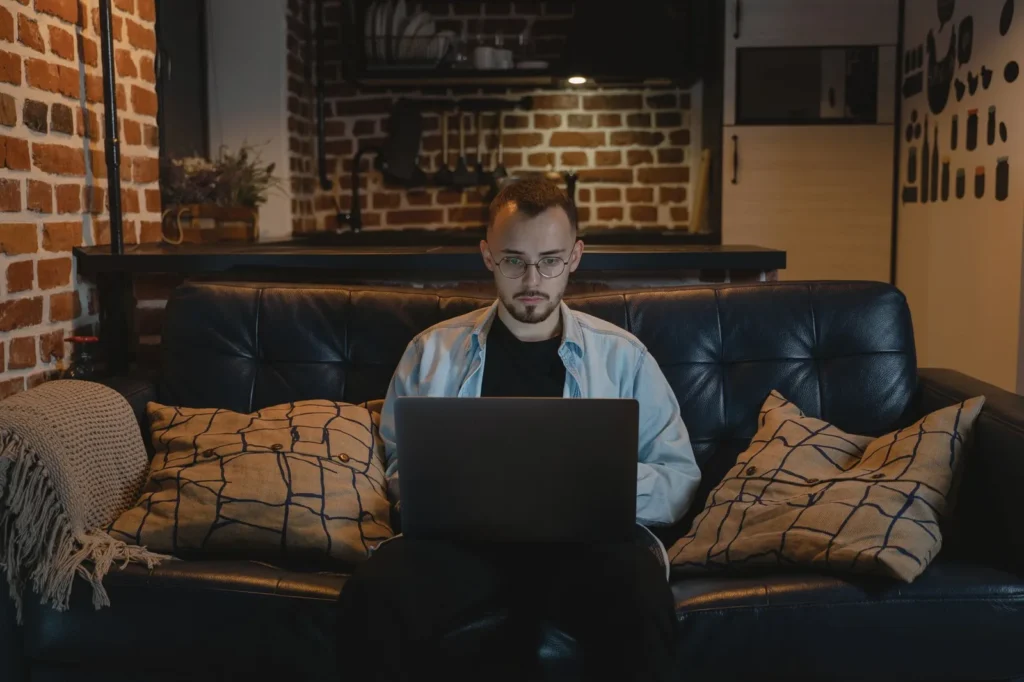 Een man werkt geconcentreerd op een laptop op een leren bank in een appartement met bakstenen muren
