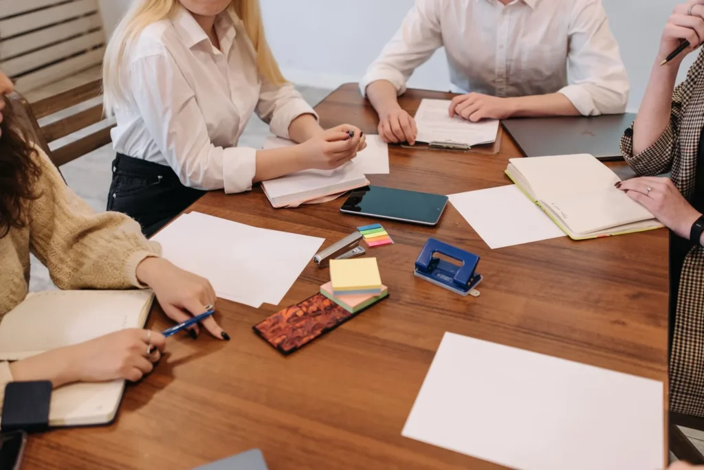 Vier mensen aan een houten tafel met notitieboeken, papieren en plakbriefjes tijdens een planningsoverleg
