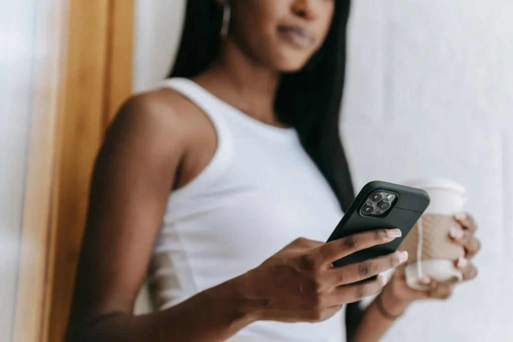 Een vrouw leunt tegen een muur en leest iets op haar smartphone, met een koffiebeker in haar andere hand