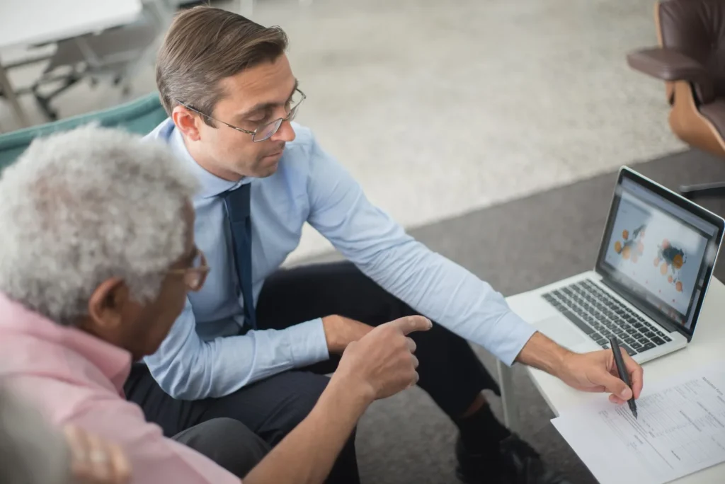 Een adviseur bespreekt een document en een laptopscherm met een oudere ondernemer aan een lage tafel