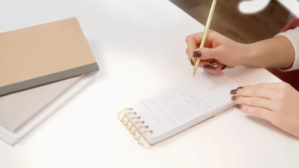 Iemand schrijft met een gouden pen aantekeningen in een klein notitieblok op een wit bureau