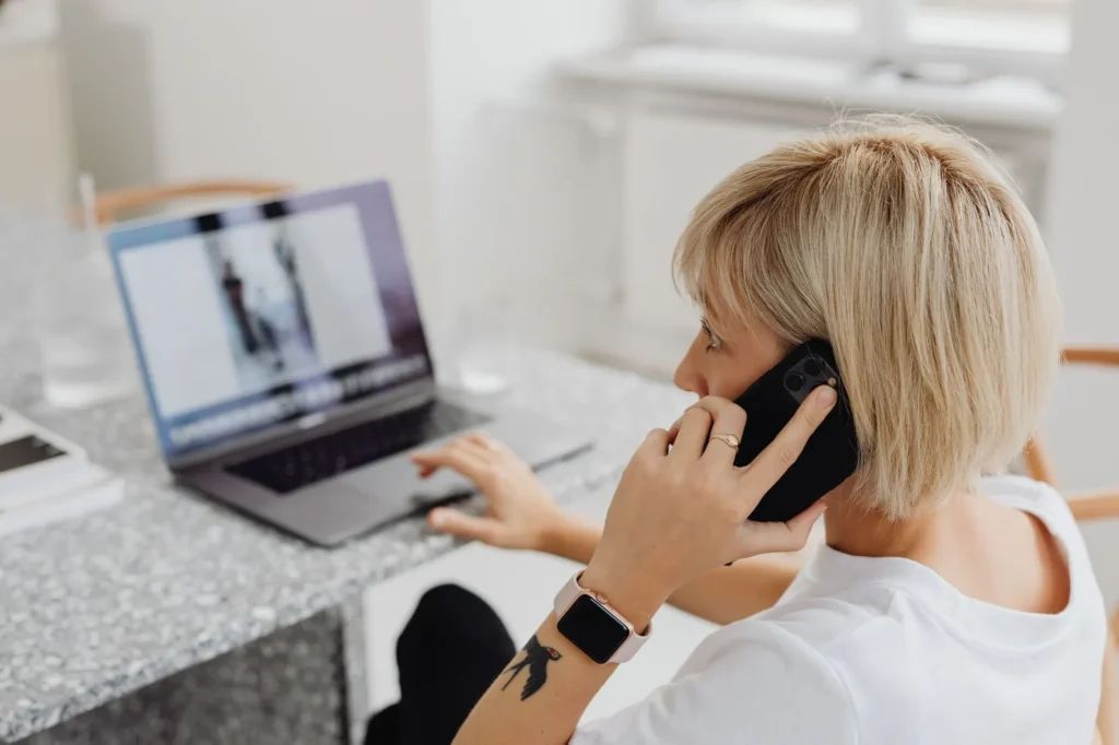 Een vrouw belt met haar telefoon terwijl ze aan een tafel op een laptop werkt