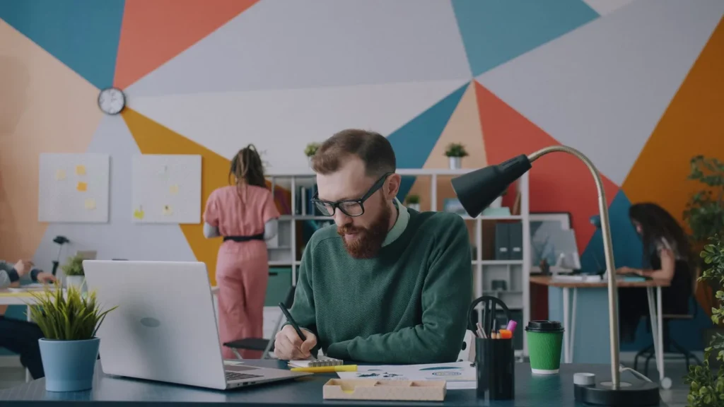 Een ontwerper met bril maakt aantekeningen naast zijn laptop in een kantoor met een kleurrijke muur, collega's werken op de achtergrond
