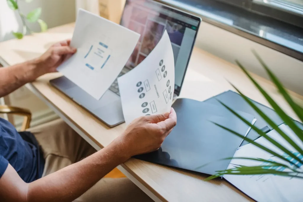 Een man houdt schetsen van webpagina's vast boven een laptop op een houten bureau